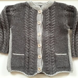 May include: A gray knitted cardigan with a cable knit pattern and wooden buttons. The cardigan has two pockets and a white trim around the neckline and edges.