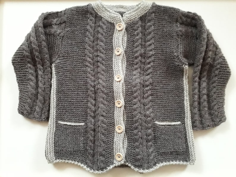 May include: A gray knitted cardigan with a cable knit pattern and wooden buttons. The cardigan has two pockets and a white trim around the neckline and edges.