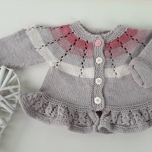 Peut inclure: Un cardigan pour bébé tricoté à la main dans les tons de gris, de rose et de blanc. Le pull présente un ourlet à volants, une fermeture à boutons et un empiècement décoratif. Les manches sont à revers et le design général est doux et délicat.