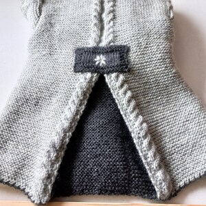 May include: A gray knitted vest with a black knitted lining and a white flower detail on the back.