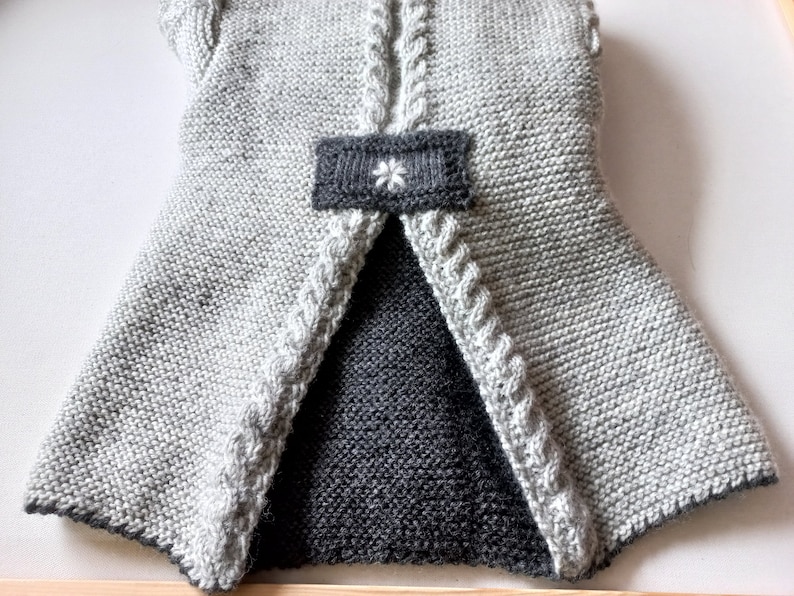 May include: A gray knitted vest with a black knitted lining and a white flower detail on the back.