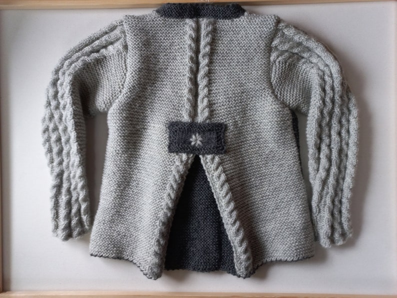 May include: A gray knitted cardigan with a gray and black flower design on the back. The cardigan has long sleeves with a cable knit pattern.