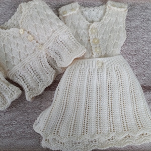 May include: A white knitted baby dress and cardigan set with a delicate lace pattern. The dress has a sleeveless bodice and a full skirt. The cardigan has long sleeves and a button closure.