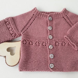 Può includere: Un cardigan per neonato di colore rosa polvere, lavorato a maglia, con chiusura a bottoni e un delicato motivo a pizzo intorno al collo e ai polsini.