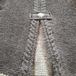 May include: Gray knitted cardigan with a white flower button on the back. The cardigan has a cable knit design on the sides and a plain knit design on the back.