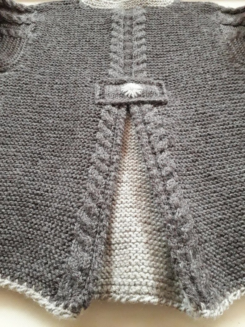 May include: Gray knitted cardigan with a white flower button on the back. The cardigan has a cable knit design on the sides and a plain knit design on the back.