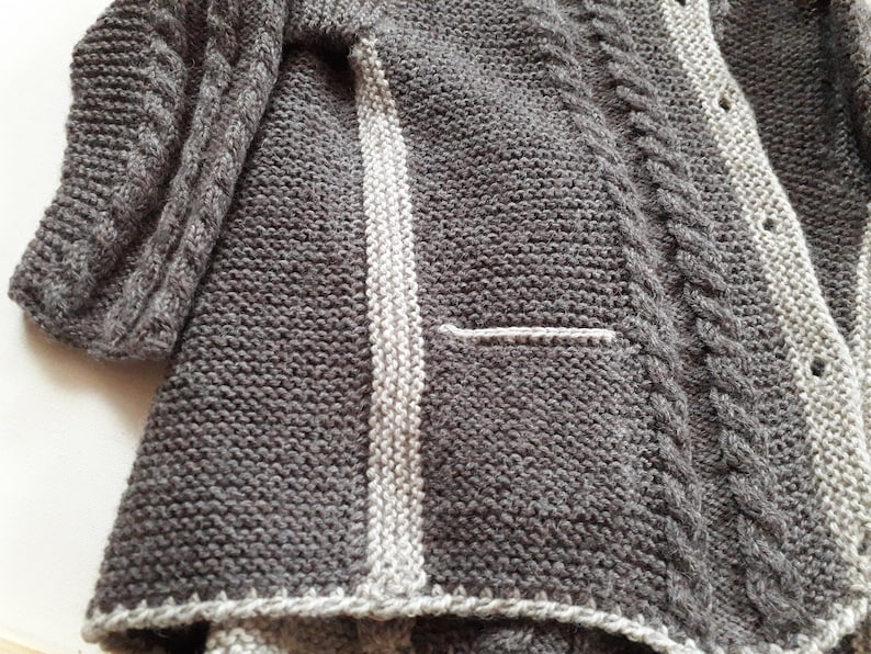 May include: A gray knitted cardigan with a white stripe down the front and a cable knit pattern on the sleeves.