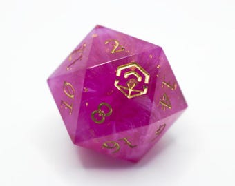 Könnte beinhalten: Ein zwanzigseitiger Würfel in Rosa und Gold. Der Würfel hat goldene Zahlen und ein goldenes Emblem. Das transparente Material hat einen Marmoreffekt mit goldenen Flocken. Ein Gaming-Zubehör.