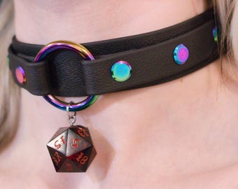 Könnte beinhalten: Schwarze Choker-Halskette mit schillernden Akzenten und einem regenbogenfarbenen Ring. Die Halskette hat einen Anhänger in Form eines zwanzigseitigen Würfels. Der Würfel ist dunkelrot mit weißen Zahlen.