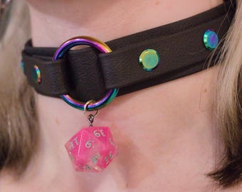 Könnte beinhalten: Schwarzes Choker-Halsband mit schillernden Akzenten. Das Halsband hat einen Regenbogenfarbenen Ring und schillernde Nieten. Ein rosa, durchscheinender, vielseitiger Würfel hängt am Ring. Ein einzigartiges Accessoire.