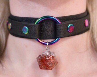 Könnte beinhalten: Schwarze Leder-Halskette mit einem großen, schillernden Metallring und einem roten und klaren Kristallanhänger. Die Halskette hat schillernde Nieten und einen passenden Ring. Der Anhänger ist ein großer, facettierter Kristall.