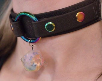 Könnte beinhalten: Ein braunes Leder-Halsband mit schillernder Hardware und Akzenten. Das Halsband hat einen großen, regenbogenfarbenen Ring und einen hängenden, vielseitigen Würfel. Das Halsband ist mit regenbogenfarbenen Nieten verziert.