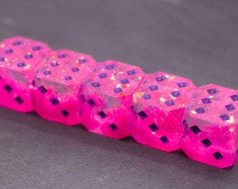 Könnte beinhalten: Fünf rosa Würfel mit lila Augen und Glitzer sind in einer Reihe angeordnet. Die Würfel haben einen Farbverlauf, der von einem dunkleren Rosa an der Basis zu einem helleren Rosa an der Oberseite übergeht. Die Würfel sind für ein Brettspiel.