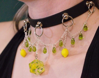 Könnte beinhalten: Schwarze Choker-Halskette mit silberfarbenen Ringen und Ketten. Die Ketten halten gelbe Zitronenanhänger, grüne Tropfenperlen und einen großen, gelben, durchscheinenden D20-Würfel. Ein einzigartiges Accessoire.