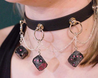 Könnte beinhalten: Schwarze Halskette mit silberfarbenen Elementen und hängenden geometrischen Anhängern. Die dunklen Anhänger haben rosa Akzente und sind mit silbernen Ketten verbunden. Die Halskette hat ein klassisches Design mit Schnallenverschluss.