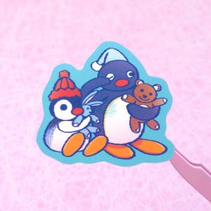 May include: A sticker featuring three cartoon penguins. One penguin wears a red hat and holds a blue bunny. Another penguin wears a blue hat and holds a brown teddy bear. The sticker has a light blue border.