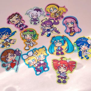 Photo of glittery stickers by marinewaltz. They are bright and colourful, and feature these synths: Hatsune Miku, Kagamine Rin and Len, Megurine Luka, SynthV Kasane Teto, Kamui Gakupo, GUMI, KAITO, MEIKO, VFlower, Big Al, Lily and Otomachi Una.
