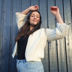 Puede incluir: Una mujer con un cárdigan de crochet blanco sobre una camisa negra y unos vaqueros azules. Está de pie frente a una pared de madera azul oscuro.