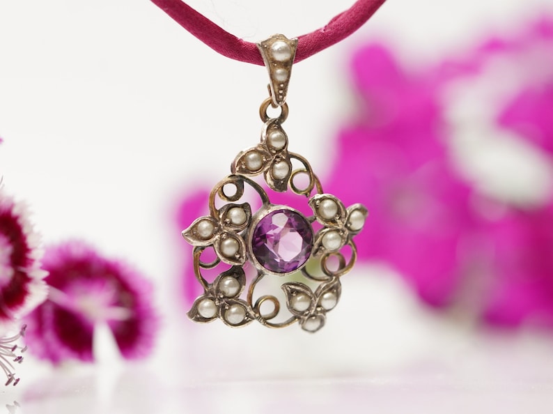 May include: A silver pendant with a purple gemstone surrounded by small pearls. The pendant is hanging from a red cord.