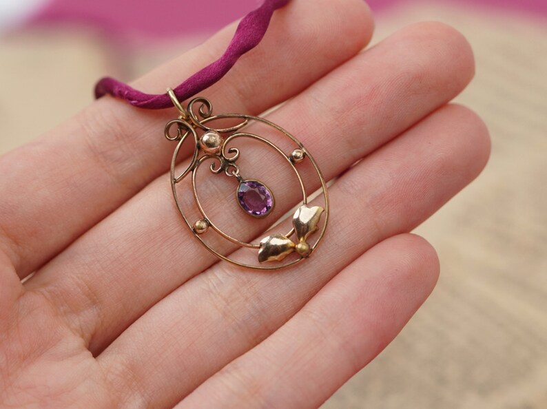 Op de afbeelding: Een delicate gouden ketting met een hanger van paarse amethist. De hanger heeft een cirkelvormig ontwerp met ingewikkelde details en een klein blaadjesmotief.