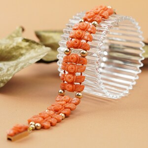 May include: A coral bracelet with carved rose motifs and gold beads. The bracelet is displayed on a clear glass stand.