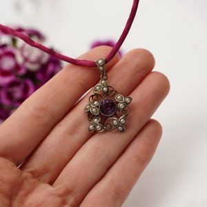 May include: A silver pendant necklace with a purple gemstone surrounded by small white pearls. The pendant is in the shape of a flower with intricate details.