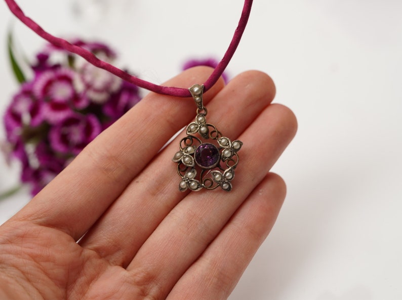 May include: A silver pendant necklace with a purple gemstone surrounded by small white pearls. The pendant is in the shape of a flower with intricate details.