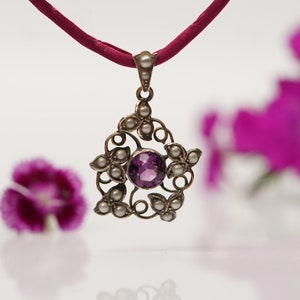 May include: A silver pendant necklace with a purple amethyst gemstone surrounded by small white pearls. The pendant is in the shape of a flower with intricate details. The necklace is on a red cord.