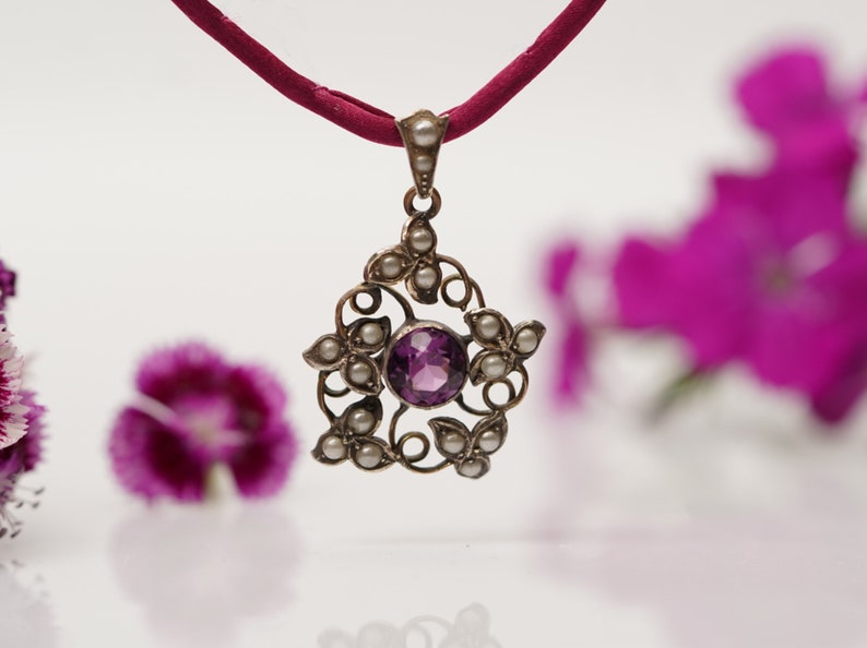 May include: A silver pendant necklace with a purple amethyst gemstone surrounded by small white pearls. The pendant is in the shape of a flower with intricate details. The necklace is on a red cord.
