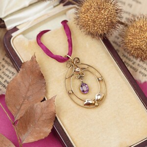 Op de afbeelding: Een delicate gouden ketting met een hanger, versierd met een paarse edelsteen en parel accenten. De hanger heeft een ovale vorm met een gedetailleerd ontwerp. De ketting wordt gepresenteerd in een fluwelen doos.