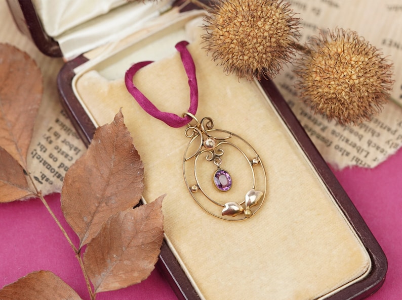 Op de afbeelding: Een delicate gouden ketting met een hanger, versierd met een paarse edelsteen en parel accenten. De hanger heeft een ovale vorm met een gedetailleerd ontwerp. De ketting wordt gepresenteerd in een fluwelen doos.