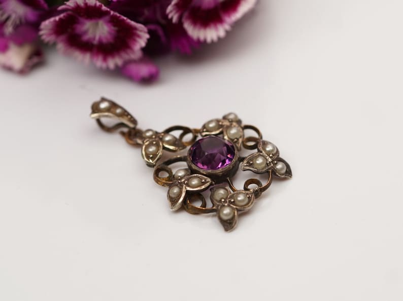 May include: A silver pendant with a purple gemstone surrounded by pearls. The pendant is in the shape of a flower with intricate details.