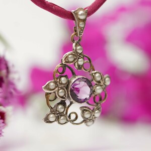 May include: A silver pendant with a purple amethyst gemstone surrounded by small white pearls. The pendant is attached to a red cord.