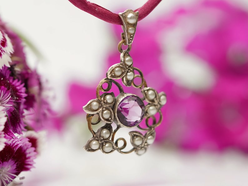 May include: A silver pendant with a purple amethyst gemstone surrounded by small white pearls. The pendant is attached to a red cord.