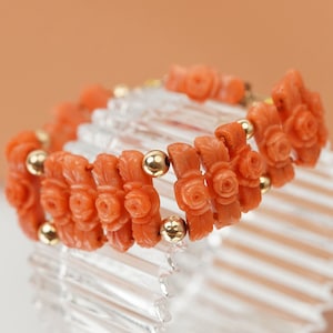 May include: A coral bracelet with carved rose motifs and gold beads. The bracelet is on a clear display stand.