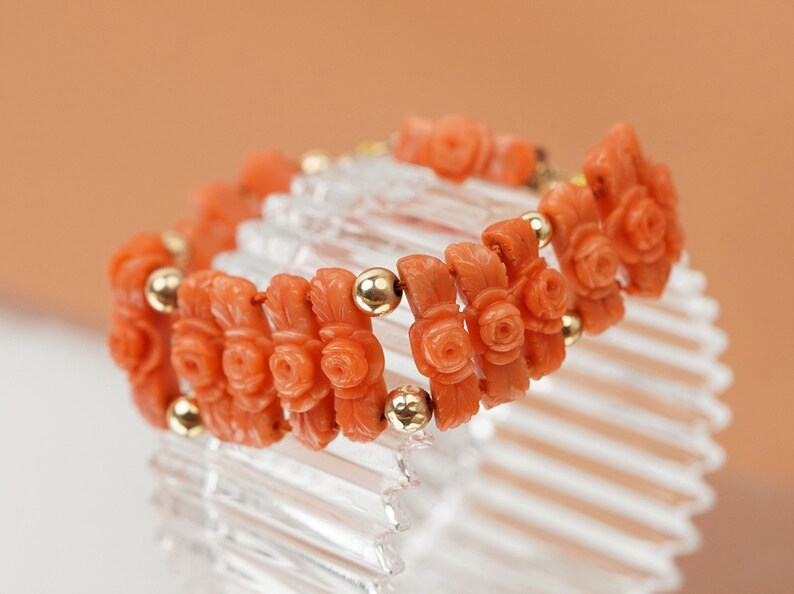 May include: A coral bracelet with carved rose motifs and gold beads. The bracelet is on a clear display stand.