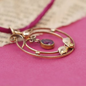 Op de afbeelding: Een gouden ketting met een hanger met een paarse edelsteen en delicate bloemmotieven. De hanger hangt aan een dun, rood koord.