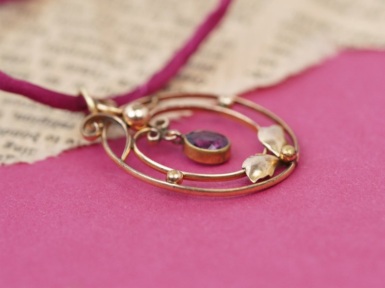Op de afbeelding: Een gouden ketting met een hanger met een paarse edelsteen en delicate bloemmotieven. De hanger hangt aan een dun, rood koord.