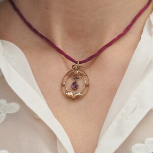 Op de afbeelding: Een delicate gouden ketting met een hanger in de vorm van een cirkel met een paarse edelsteen. De ketting is aan een donkerrode koord.