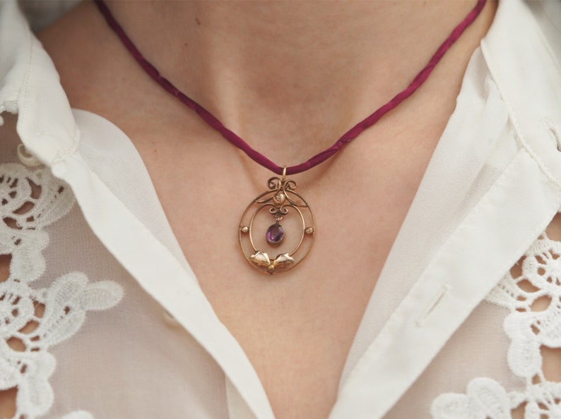 Op de afbeelding: Een delicate gouden ketting met een hanger in de vorm van een cirkel met een paarse edelsteen. De ketting is aan een donkerrode koord.