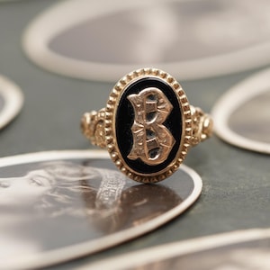 May include: A vintage gold ring with an oval black onyx stone. The stone features a raised gold letter "B". The ring has a detailed gold band with decorative elements. The ring is set against a backdrop of vintage photographs.