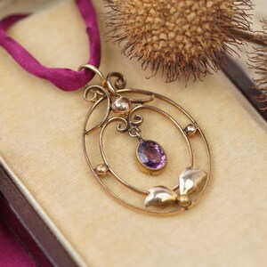 Op de afbeelding: Een gouden ketting met een hanger met een paarse edelsteen in een delicate, sierlijke setting. De hanger hangt aan een dunne gouden ketting en aan een rood lint.