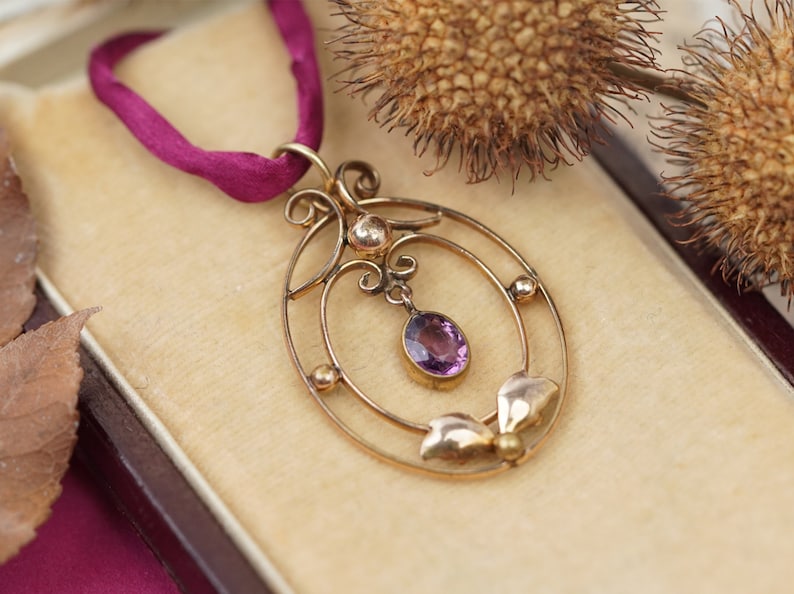 Op de afbeelding: Een gouden ketting met een hanger met een paarse edelsteen in een delicate, sierlijke setting. De hanger hangt aan een dunne gouden ketting en aan een rood lint.