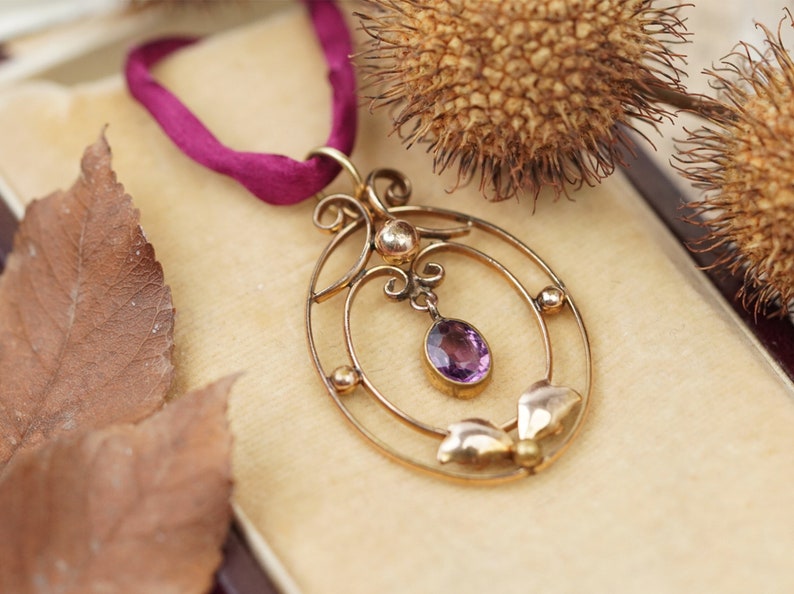 Op de afbeelding: Een delicate gouden ketting met een hanger van paarse amethist. De hanger heeft een cirkelvormig ontwerp met ingewikkelde details en een hangende amethistdruppel.