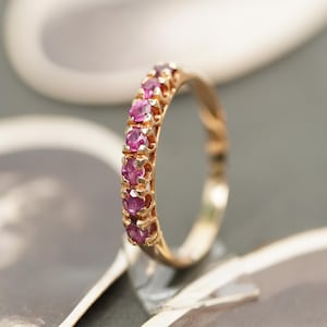 Op de afbeelding: Een gouden ring met een rij roze edelstenen. De ring is bezet met meerdere kleine, ronde edelstenen. De ring is gemaakt van goud en heeft een gepolijste afwerking. De ring is een sieraad.