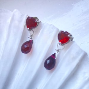May include: A pair of silver earrings with red heart-shaped stones and teardrop-shaped stones hanging below.