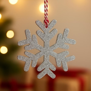 Puede incluir: Adorno navideño de copo de nieve plateado con purpurina, colgado de una cuerda roja y blanca. El copo de nieve tiene seis puntas y está suspendido sobre un fondo borroso con luces cálidas y regalos envueltos.