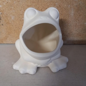 May include: A ceramic frog-shaped container, unpainted and ready for customization. The frog has large, round eyes and an open mouth, forming a bowl. The frog is sitting on its feet, with a neutral background.