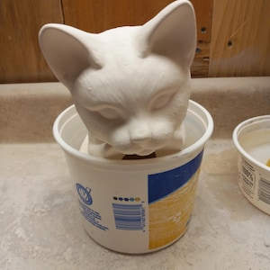 May include: A white ceramic cat head figurine sits inside a white and blue plastic container. The cat has pointed ears and closed eyes. The container has a yellow and blue design and a barcode.