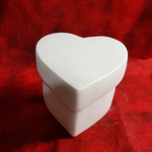May include: A white heart-shaped box with a matching lid. The box is sitting on a red surface. The box is a solid color and has a smooth finish. The box is likely used for storing small items or as a decorative piece.